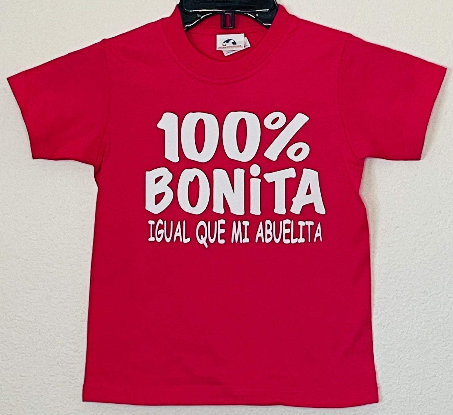 "100% Bonita Cómo Mi Abuelita" Youth Silkscreen T-Shirt in hot pink with bold black text, showcasing a playful and heartwarming design. Displayed flat with a clean background, available in sizes XS-XL. Perfect for kids who love their grandma!