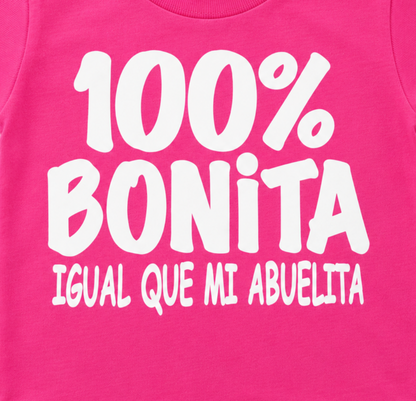 Close-up of the 100% Bonita Igual Que Mi Abuelita infant shirt design with bold white lettering on hot pink.
