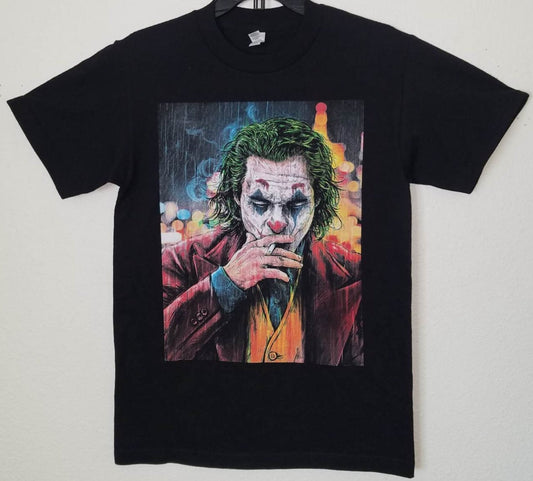"2019 Joker Smoking T-Shirt featuring a colorful graphic of the Joker in makeup and suit, smoking against a city lights backdrop. Black unisex tee, available in sizes Small to 3XL."