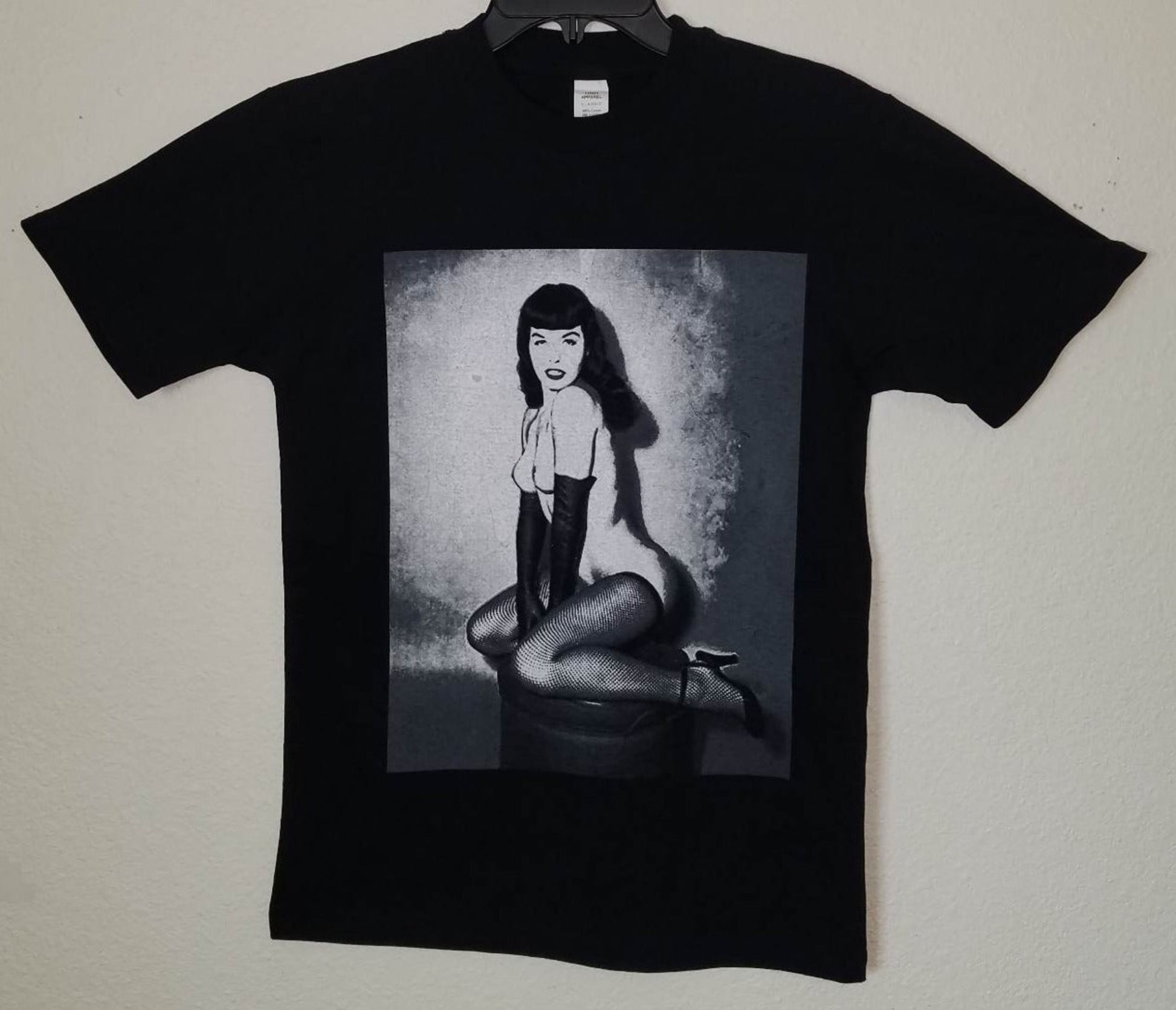 Black unisex “Bettie Page – On Her Knees” T-shirt featuring grayscale vintage pin-up artwork with retro photo style, available in sizes Small–3XL.