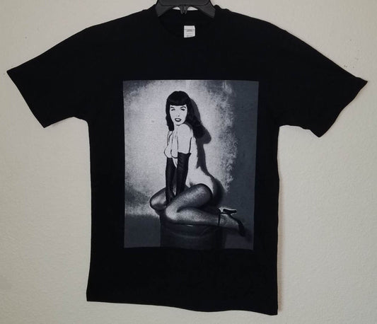 Black unisex “Bettie Page – On Her Knees” T-shirt featuring grayscale vintage pin-up artwork with retro photo style, available in sizes Small–3XL.