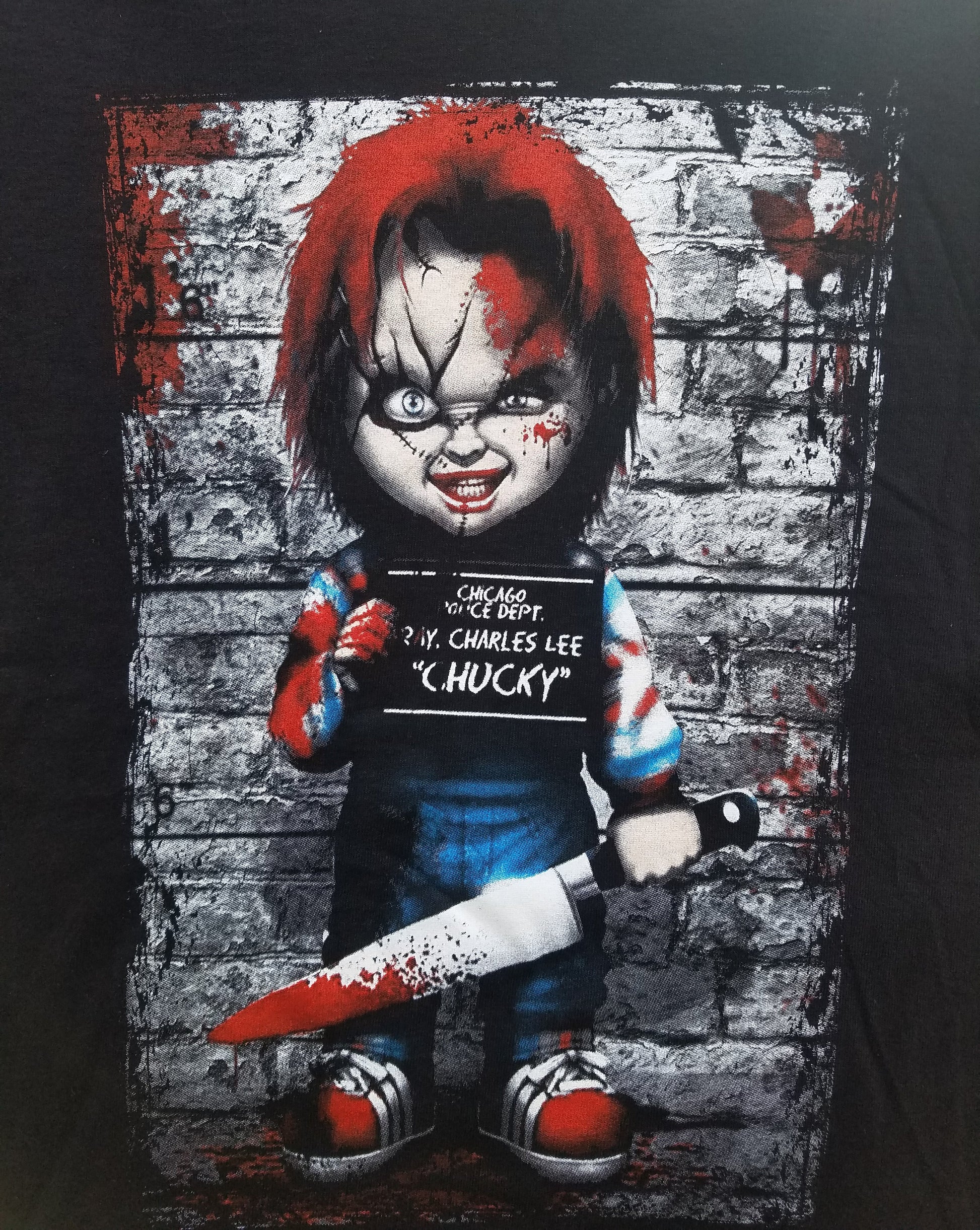 Front view of Chucky Mugshot T-shirt featuring horror doll graphic with bloody knife and mugshot background printed in authentic silkscreen on black cotton tee.