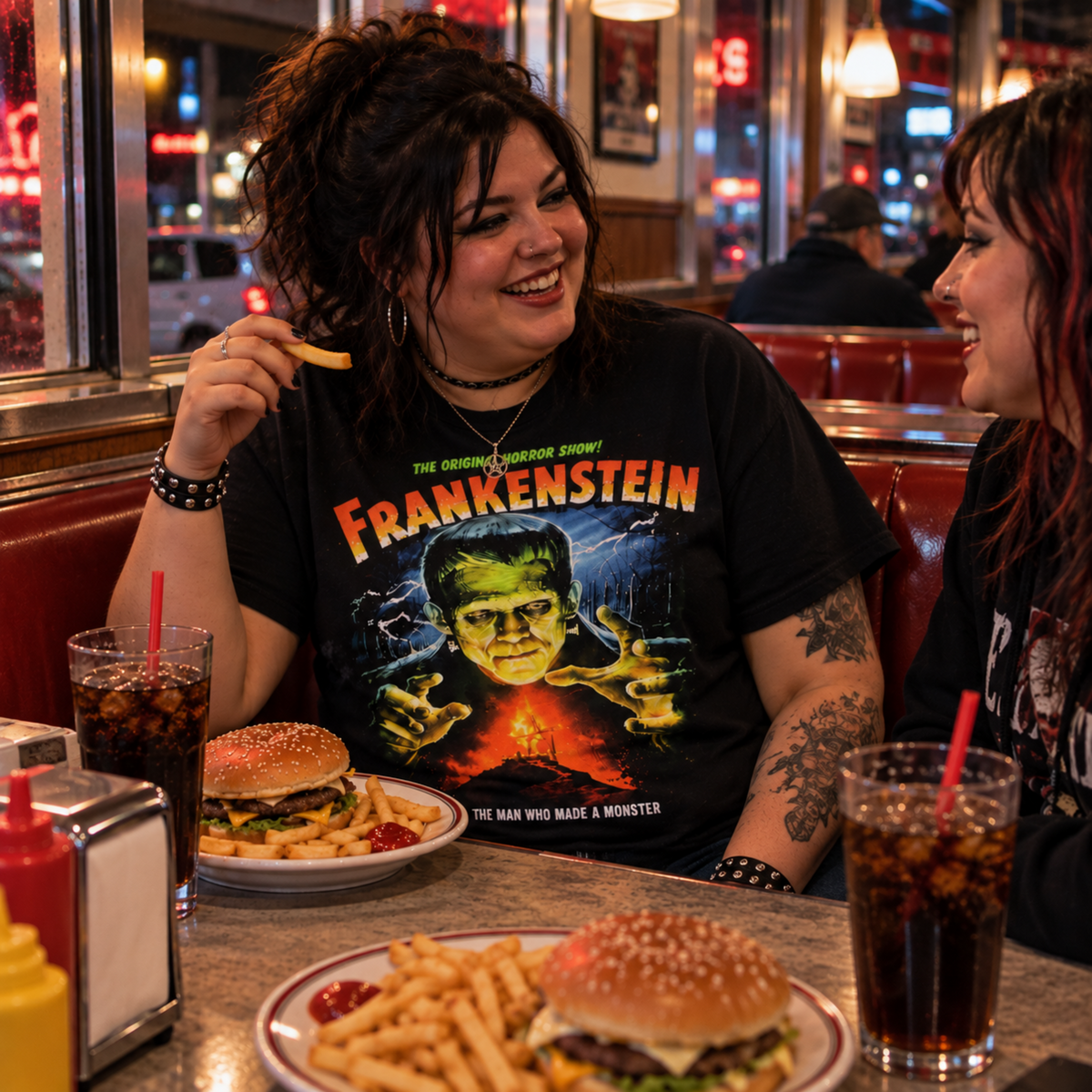 Woman wearing the black Frankenstein T-shirt while eating burgers and fries with a friend at a diner—a strong pick for classic monster and horror fans.