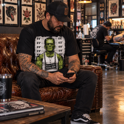 Man wearing the Frankenstein Monster Mugshot black T-shirt while seated inside a tattoo shop, featuring the green monster, height chart and prisoner placard.