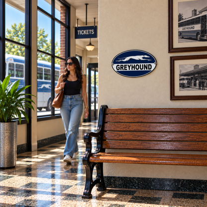 Greyhound Bus Station oval metal sign displayed above a wooden bench in a vintage-style bus terminal waiting area with historic bus photographs.