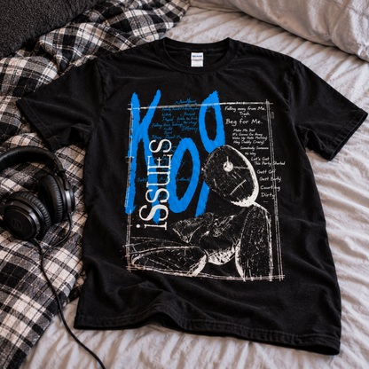 Black Issues graphic T-shirt displayed on a bed with headphones and flannel nearby, featuring the large blue ISSUES lettering and illustrated front design.
