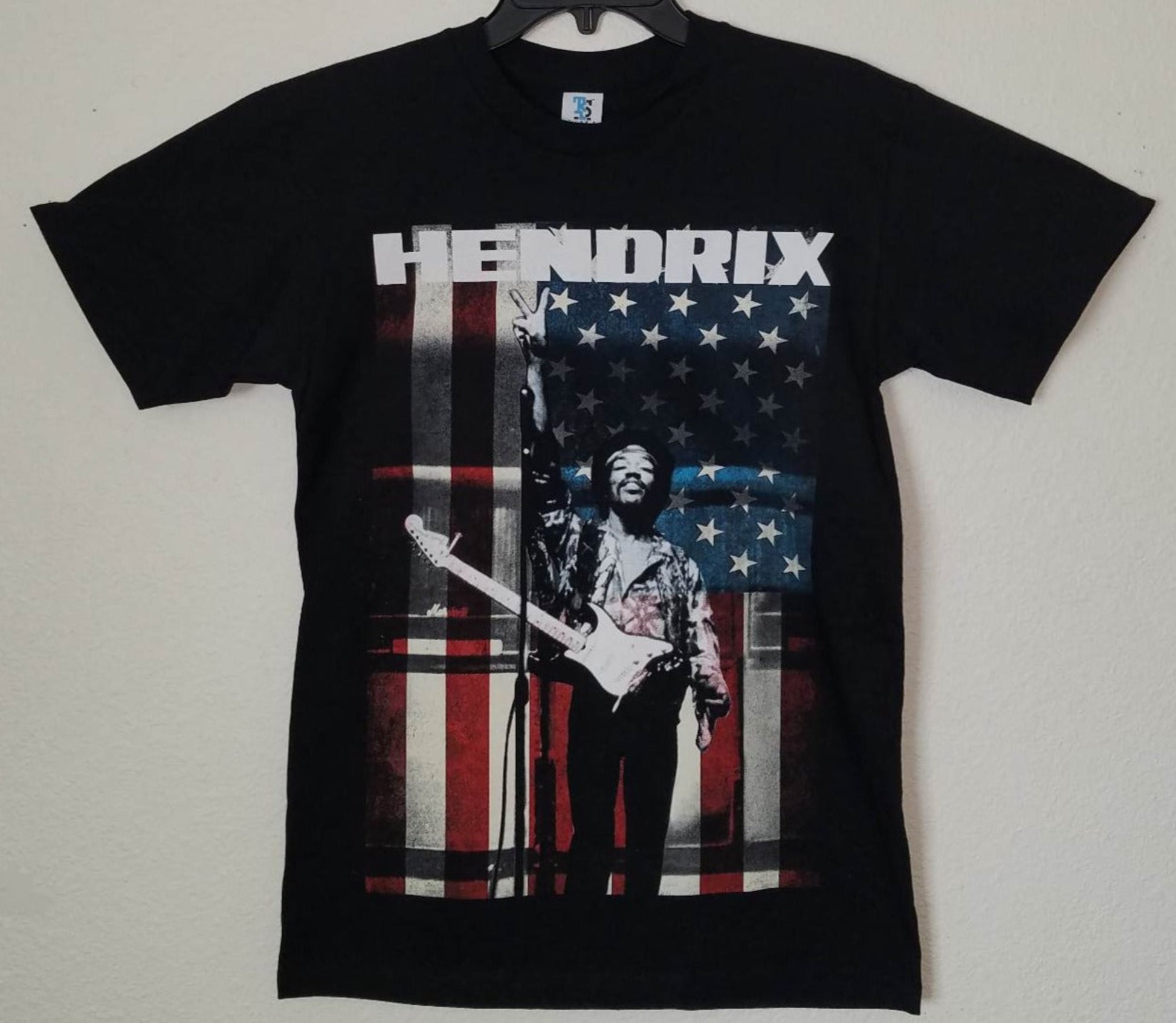 Black “Jimi Hendrix – Peace Sign” unisex T-shirt featuring Hendrix standing before an American flag with guitar in hand, Small–3XL.
