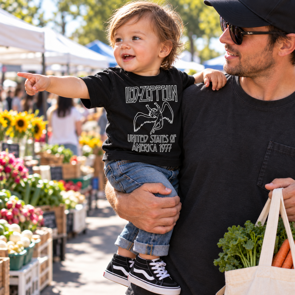 Child wearing a Led Zeppelin America 1977 black infant graphic T-shirt while being held by Dad at an outdoor farmers market.