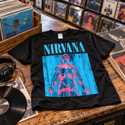 Nirvana Sliver black T-shirt displayed on a record store counter surrounded by vinyl, featuring the bold blue and pink graphic—a strong pick for grunge fans.