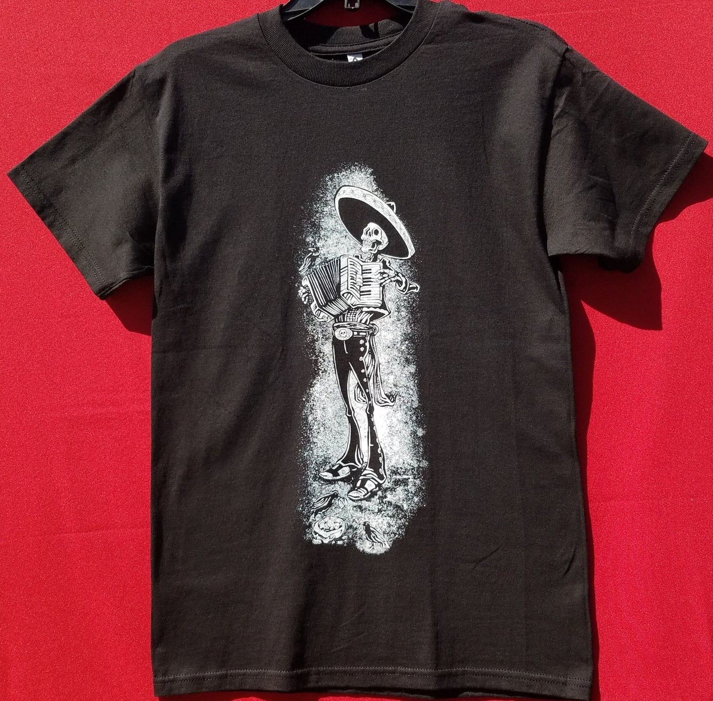 Skeleton Mariachi Accordion Player T-Shirt – A detailed graphic of a skeleton dressed in traditional mariachi attire, passionately playing an accordion. Bold and eye-catching design on a high-quality cotton tee. Available in sizes Small-2XL.