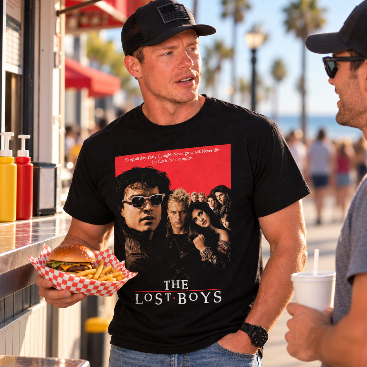 Man wearing The Lost Boys black graphic T-shirt while holding a burger basket at a boardwalk food stand, featuring the original 1987 movie-cover design.