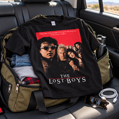 The Lost Boys black graphic T-shirt displayed with packed road-trip gear in a car, featuring the original 1987 movie-cover artwork and cast imagery.