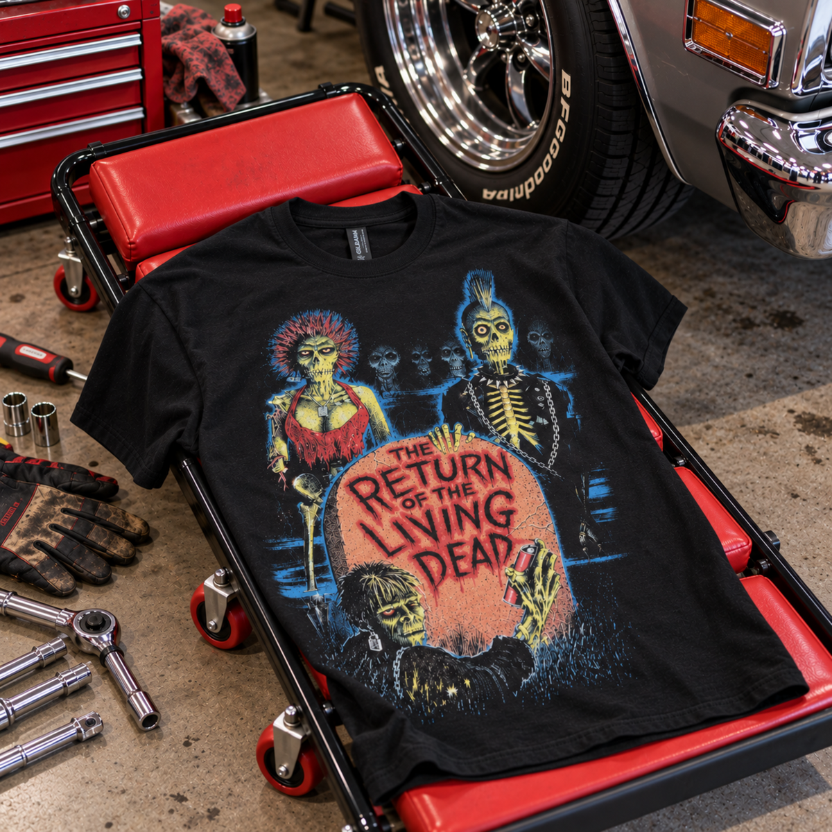 The Return of the Living Dead 1985 black graphic T-shirt displayed on a garage creeper beside tools and a classic car, featuring the colorful zombie tombstone design.